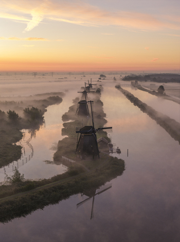Aerial view of UNESCO World Heritage Kinderdijk windmills emerging from thick morning mist along the canals under a golden sunrise sky Kinderdijk Zuid Holland Netherlands.