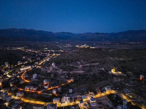 Aerial view of Saranda at night with illuminated streets winding through the hillside buildings glowing and distant mountains under a deep blue sky in Saranda Vlore County Albania. Aerial view of Saranda at night with illuminated streets winding through the hillside buildings glowing and distant mountains under a deep blue sky in Saranda Vlore County Albania.