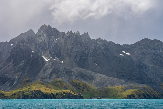 View of the rugged peaks and green slopes of Drygalski Fjord rising above the turquoise waters under a cloudy sky in South Georgia and the South Sandwich Islands.