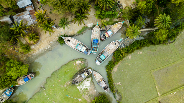 Aerial view of wooden fishing boats moored in a narrow canal surrounded by lush green vegetation and agricultural fields in Patharghata Barisal Division Bangladesh. Aerial view of wooden fishing boats moored in a narrow canal surrounded by lush green vegetation and agricultural fields in Patharghata Barisal Division Bangladesh.