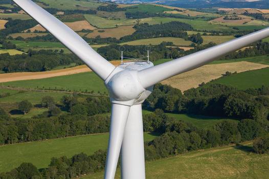 Aerial view of a colossal wind turbine its white blades slicing through the blue sky above the patchwork fields contrasting with the lush green and brown landscape Salles Curan Occitanie France. Aerial view of a colossal wind turbine its white blades slicing through the blue sky above the patchwork fields contrasting with the lush green and brown landscape Salles Curan Occitanie France.