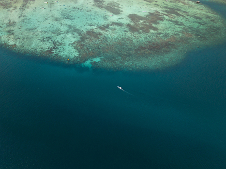 Aerial view of a solitary boat glides across the tranquil deep blue sea contrasting with the vibrant turquoise reef below Karimunjawa Central Java Indonesia. Aerial view of a solitary boat glides across the tranquil deep blue sea contrasting with the vibrant turquoise reef below Karimunjawa Central Java Indonesia.