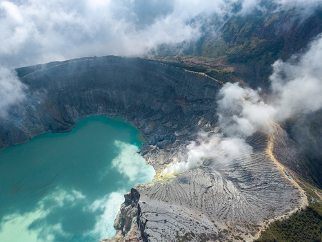 Aerial view of Ijen crater lake with its vibrant turquoise water and sulfurous volcanic smoke rising from the rugged rocky rim in Ijen East Java Indonesia.