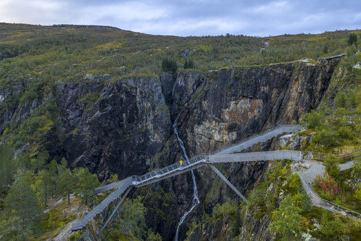 Aerial view of Voringfossen waterfall and the modern staircase bridge spanning the deep rocky canyon with a person in a yellow jacket Eidfjord Municipality Vestland Norway.