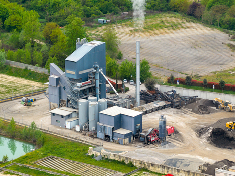 Aerial view of an industrial asphalt mixing plant with tall chimneys emitting smoke storage silos and heavy machinery surrounded by green fields in San Giovanni Valdarno Tuscany Italy.