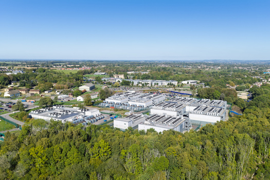 Aerial view of a data center facility surrounded by lush green trees and vegetation with parking areas and nearby buildings in the United Kingdom.
