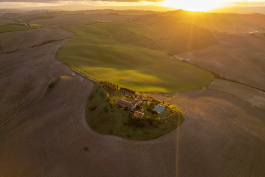 Aerial view of a farmhouse with a swimming pool and cypress trees set in the rolling hills of the Crete Senesi landscape during golden hour in Asciano Tuscany Italy.