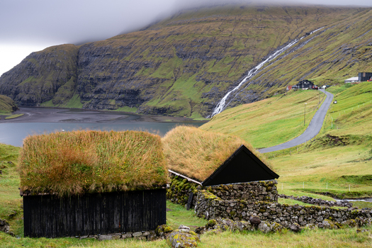 View of traditional grass roofed houses in the foreground with a waterfall cascading down a lush green mountain slope and a winding road leading to a fjord in the Faroe Islands.