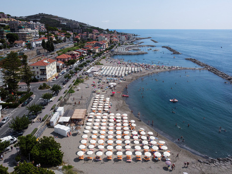 Aerial view of the sandy beach with rows of white umbrellas and people swimming in the blue sea along the coastal town San Lorenzo Liguria Italy.