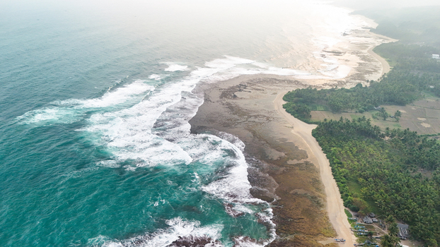 Aerial view of a rugged coastline with turquoise waves crashing against the rocky shore bordered by a lush palm tree forest and sandy beach in Indonesia. Aerial view of a rugged coastline with turquoise waves crashing against the rocky shore bordered by a lush palm tree forest and sandy beach in Indonesia.