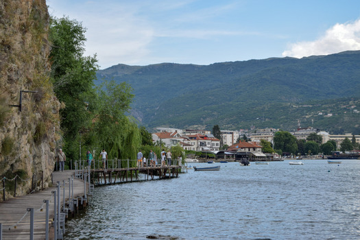 View of the wooden boardwalk along Lake Ohrid with people walking rocky cliffs and mountains in the background in Ohrid Municipality of Ohrid North Macedonia. View of the wooden boardwalk along Lake Ohrid with people walking rocky cliffs and mountains in the background in Ohrid Municipality of Ohrid North Macedonia.