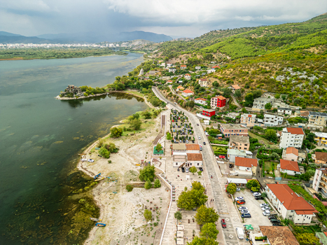 Aerial view of the lakeside village featuring a promenade parked cars houses with red roofs and the calm waters of Lake Shkodra. Shiroka Shkoder County Albania.