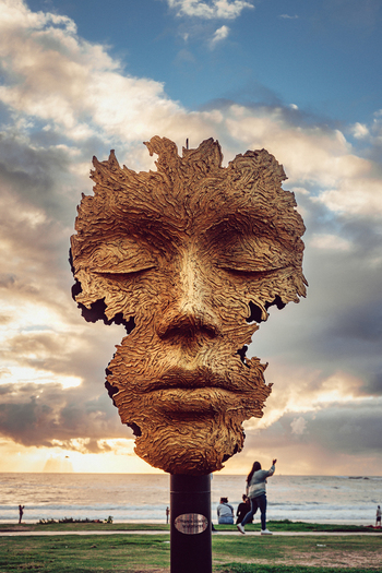 View of a large textured face sculpture with closed eyes on the Sea Point Promenade at sunset with people and the ocean in Cape Town Western Cape South Africa. View of a large textured face sculpture with closed eyes on the Sea Point Promenade at sunset with people and the ocean in Cape Town Western Cape South Africa.