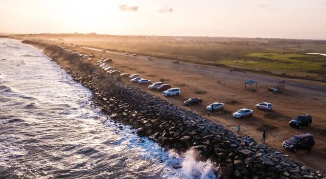 Aerial view of a coastal road lined with parked cars along a rocky breakwater with ocean waves crashing against the shore during sunset in Ghana. Aerial view of a coastal road lined with parked cars along a rocky breakwater with ocean waves crashing against the shore during sunset in Ghana.