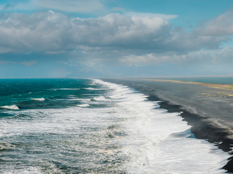 Aerial view of Stokksnes beach with its iconic black sand crashing white waves and dramatic mountains under a cloudy sky in Iceland.