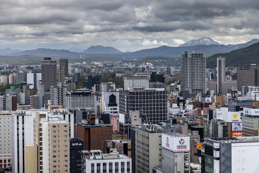 Aerial view of the dense urban skyline of Sapporo featuring modern skyscrapers and high rise buildings set against a backdrop of distant mountains under a cloudy sky in Sapporo Hokkaido Japan. Aerial view of the dense urban skyline of Sapporo featuring modern skyscrapers and high rise buildings set against a backdrop of distant mountains under a cloudy sky in Sapporo Hokkaido Japan.