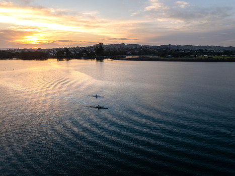 Raglan New Zealand 09 February 2026 Aerial view of two rowers gliding across the calm waters of the bay during a golden sunset with the coastal town visible in the distance.