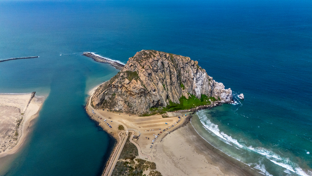 Aerial view of Morro Rock standing prominently on the coastline with sandy beaches a parking area and the blue waters of the Pacific Ocean in Morro Bay California United States.