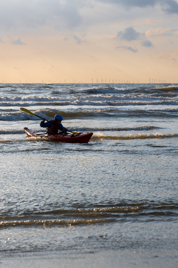 View of a person in a red kayak paddling through the North Sea waves with wind turbines on the horizon at Strand Wijk aan Zee North Holland Netherlands. View of a person in a red kayak paddling through the North Sea waves with wind turbines on the horizon at Strand Wijk aan Zee North Holland Netherlands.
