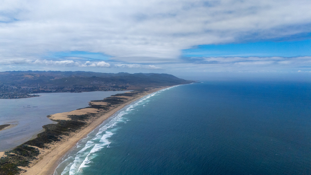 Aerial view of the long sandspit separating the calm bay from the Pacific Ocean with crashing waves and distant hills in Morro Bay California United States.