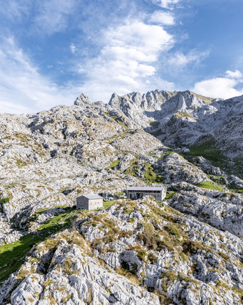 Aerial view of stone refuge nestled amidst rugged sun kissed peaks where the sharp grey rock meets the soft green meadows Picos de Europa Asturias Spain.