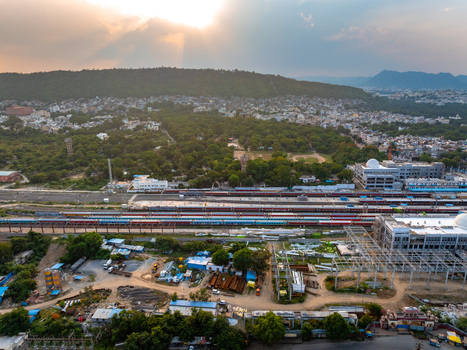 Aerial view of the railway station with trains on tracks surrounded by urban buildings lush trees and hills under a warm sunset sky in Udaipur Rajasthan India. Aerial view of the railway station with trains on tracks surrounded by urban buildings lush trees and hills under a warm sunset sky in Udaipur Rajasthan India.
