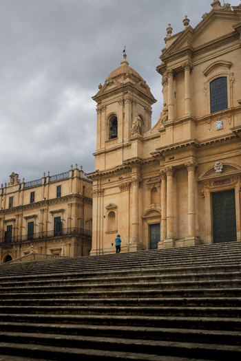 View of the baroque facade of Noto Cathedral with its grand stone staircase and a person standing on the steps under a cloudy sky in Noto Sicily Italy. View of the baroque facade of Noto Cathedral with its grand stone staircase and a person standing on the steps under a cloudy sky in Noto Sicily Italy.
