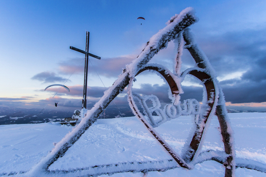 View of a snow covered cross and heart shaped structure with paragliders soaring in the distance against a twilight sky Schöckl Styria Austria. View of a snow covered cross and heart shaped structure with paragliders soaring in the distance against a twilight sky Schöckl Styria Austria.