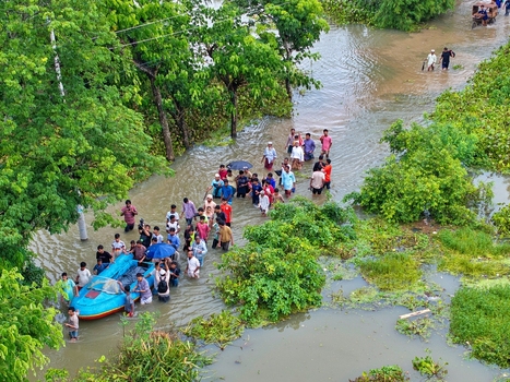 Banshkhali Upazila Bangladesh 10 July 2026 Aerial view of people wading through floodwaters in a submerged area with a small blue boat being guided by locals amidst lush green trees. Banshkhali Upazila Bangladesh 10 July 2026 Aerial view of people wading through floodwaters in a submerged area with a small blue boat being guided by locals amidst lush green trees.