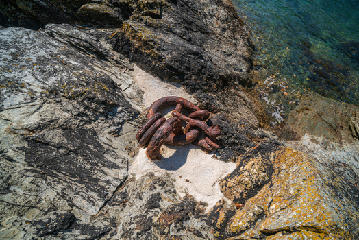 View of a heavy rusty metal chain resting on jagged coastal rocks beside the clear water of the Fowey River in Fowey Cornwall United Kingdom. View of a heavy rusty metal chain resting on jagged coastal rocks beside the clear water of the Fowey River in Fowey Cornwall United Kingdom.
