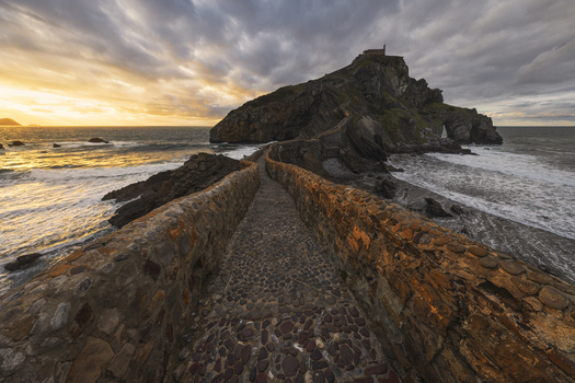 View of the stone bridge leading to the hermitage on a rocky island under a dramatic sunset sky with crashing waves Gaztelugatxeko Doniene Euskadi Spain. View of the stone bridge leading to the hermitage on a rocky island under a dramatic sunset sky with crashing waves Gaztelugatxeko Doniene Euskadi Spain.