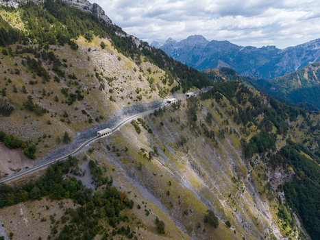 Aerial view of the winding mountain road with tunnels carved into the rocky slopes of the Albanian Alps surrounded by pine forests and rugged terrain in Shkoder Shkoder County Albania. Aerial view of the winding mountain road with tunnels carved into the rocky slopes of the Albanian Alps surrounded by pine forests and rugged terrain in Shkoder Shkoder County Albania.
