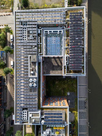 Aerial view of a modern building featuring extensive rooftop solar panels a central glass roofed courtyard and green terraces situated along a river in Germany.