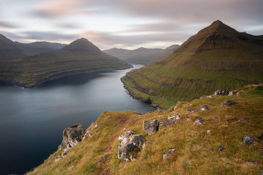 View of the dramatic landscape of the Faroe Islands with steep green mountains a deep fjord and rocky foreground under a soft cloudy sky Faroe Islands. View of the dramatic landscape of the Faroe Islands with steep green mountains a deep fjord and rocky foreground under a soft cloudy sky Faroe Islands.