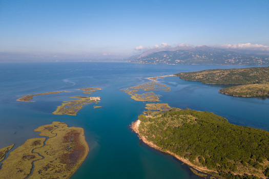 Aerial view of Kanali i Vivarit with lush green hills and wetlands along the calm blue water under a clear sky in Vrine Vlore County Albania. Aerial view of Kanali i Vivarit with lush green hills and wetlands along the calm blue water under a clear sky in Vrine Vlore County Albania.