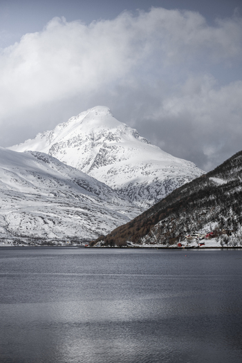 View of snow covered mountains and a calm fjord with small red cabins along the shoreline under a cloudy sky Kvaloya Troms Norway. View of snow covered mountains and a calm fjord with small red cabins along the shoreline under a cloudy sky Kvaloya Troms Norway.