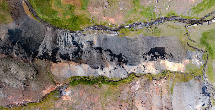 Aerial view of the rugged volcanic terrain of Grensdalur featuring dark basaltic rock geothermal soil and winding streams surrounded by vibrant green moss in Sveitarfelagid Olfus Iceland. Aerial view of the rugged volcanic terrain of Grensdalur featuring dark basaltic rock geothermal soil and winding streams surrounded by vibrant green moss in Sveitarfelagid Olfus Iceland.