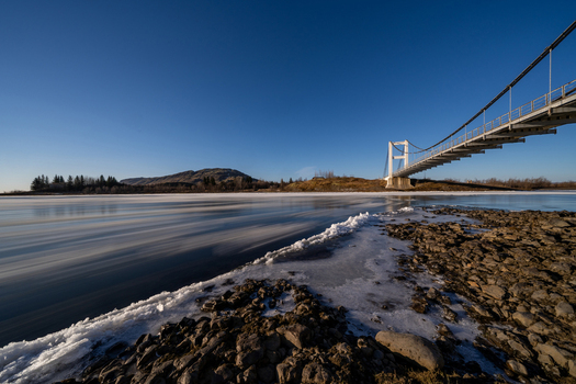 View of Laugaras Bridge spanning a frozen river with ice floes and rocky banks under a clear blue sky in Blaskogabyggd Iceland. View of Laugaras Bridge spanning a frozen river with ice floes and rocky banks under a clear blue sky in Blaskogabyggd Iceland.