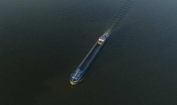 Aerial view of a long cargo barge navigating dark river water with a visible wake during golden hour in Netherlands. Aerial view of a long cargo barge navigating dark river water with a visible wake during golden hour in Netherlands.