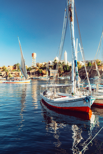 View of traditional felucca sailboats moored on the calm blue waters of the Nile River with historic buildings and palm trees along the shore in Aswan Aswan Governorate Egypt. View of traditional felucca sailboats moored on the calm blue waters of the Nile River with historic buildings and palm trees along the shore in Aswan Aswan Governorate Egypt.
