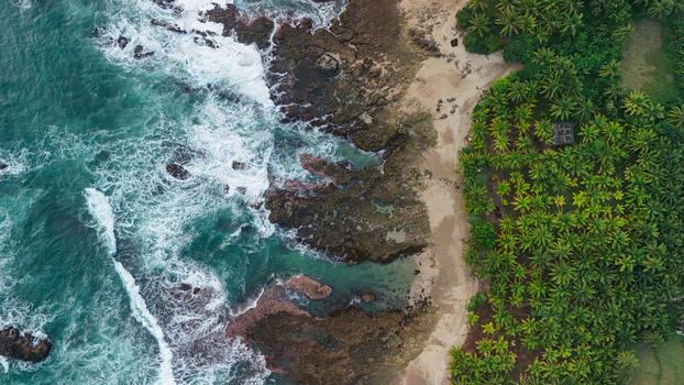 Aerial view of a tropical beach with turquoise waves crashing against rocky cliffs and a dense palm tree forest along the coastline in Indonesia. Aerial view of a tropical beach with turquoise waves crashing against rocky cliffs and a dense palm tree forest along the coastline in Indonesia.
