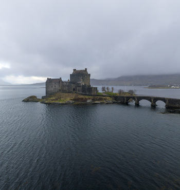 Aerial view of Eilean Donan Castle on a small island connected by a stone bridge over Loch Duich under a misty sky in Dornie Scotland United Kingdom.