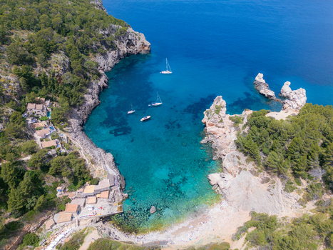 Aerial view of Cala Deia with turquoise waters sailboats anchored in the bay and rocky cliffs covered in lush pine trees in Spain. Aerial view of Cala Deia with turquoise waters sailboats anchored in the bay and rocky cliffs covered in lush pine trees in Spain.