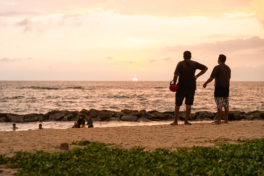 Sri Lanka 14 January 2026 View of two men standing on a sandy beach watching the sunset over the Indian Ocean with people swimming near the shore.