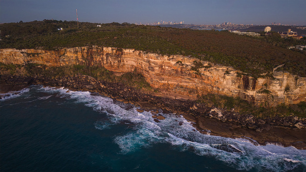 Aerial view of the rugged sandstone cliffs of North Head meeting the Pacific Ocean with waves crashing against the rocky shoreline in Manly New South Wales Australia.