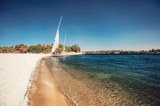 View of a traditional felucca sailboat anchored near a sandy beach on the Nile River with palm trees and clear blue sky in Aswan Aswan Governorate Egypt. View of a traditional felucca sailboat anchored near a sandy beach on the Nile River with palm trees and clear blue sky in Aswan Aswan Governorate Egypt.
