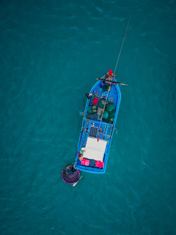 Aerial view of a small blue fishing boat with a round basket boat attached carrying fishing nets and a solar panel on the deck floating in turquoise sea water in Vietnam. Aerial view of a small blue fishing boat with a round basket boat attached carrying fishing nets and a solar panel on the deck floating in turquoise sea water in Vietnam.