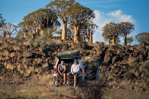 Quiver Tree Forest Namibia 06 March 2026 View of two individuals resting on a vehicle s open tailgate amidst the unique Quiver Trees and rugged rocky terrain. Quiver Tree Forest Namibia 06 March 2026 View of two individuals resting on a vehicle s open tailgate amidst the unique Quiver Trees and rugged rocky terrain.