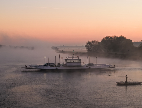 Aerial view of a car ferry crossing a misty river during a soft orange sunrise with thick fog rolling over the water and distant trees Kessel Limburg Netherlands.