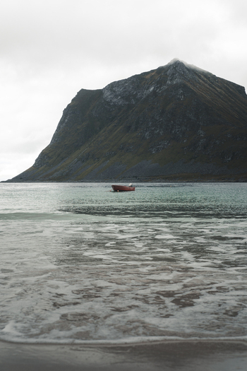 View of a small red boat floating in the turquoise water of Haukland Beach with a large mountain rising in the background under a cloudy sky in Leknes Nordland Norway. View of a small red boat floating in the turquoise water of Haukland Beach with a large mountain rising in the background under a cloudy sky in Leknes Nordland Norway.